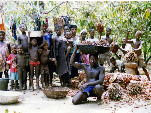 A group of children and adults gather in a lush outdoor setting, some carrying baskets of reddish fruits, with clothes hung on trees